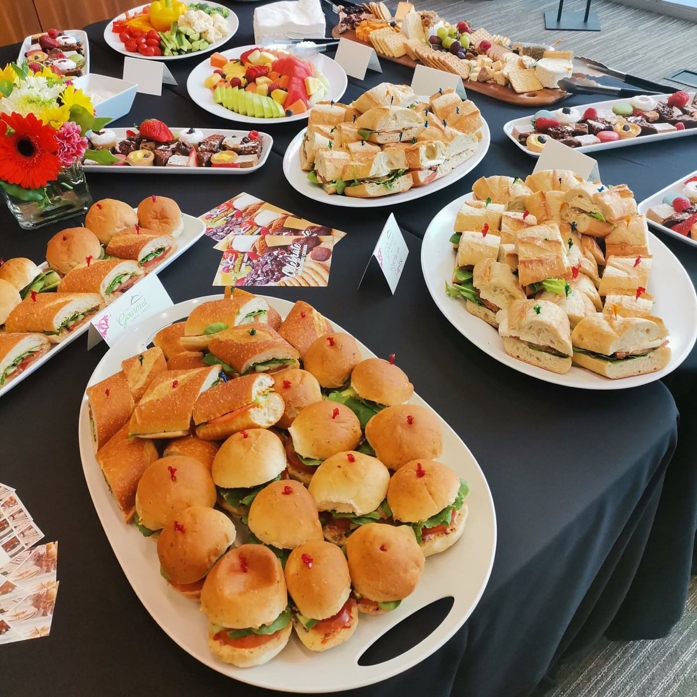 A catering buffet spread of sandwich platters, a cheese and cracker board, fruit platters, and dessert trays, set up in a reception room